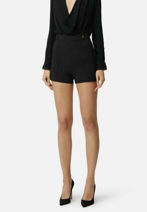 Donna con pantaloncini neri a vita alta, top nero a maniche lunghe con scollo a V e tacchi alti neri con punta a punta, in piedi davanti a uno sfondo bianco.