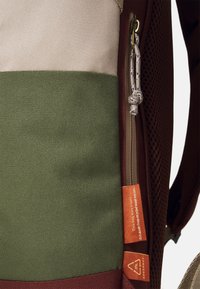 Reppu, jossa on väri-blokki design, jossa on beige, vihreä ja marooni. Materiaaleina on teksturoitu kangas ja verkko. Säädettävä vetoketjun oikea puoli.