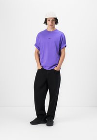 Purple cotton t-shirt with rolled sleeves, black loose-fitting trousers, and black sneakers. Model wears a white cap with logo.