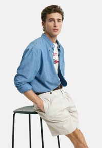 Camisa de botones azul claro, camiseta gráfica blanca debajo, pantalones cortos beige, cinturón oscuro, sentado en un taburete de metal negro frente a un fondo blanco.