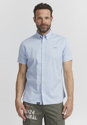 Camisa de botón de manga corta azul claro hecha de algodón, con un pequeño diseño bordado en la bolsa del pecho con acentos en rojo y azul.