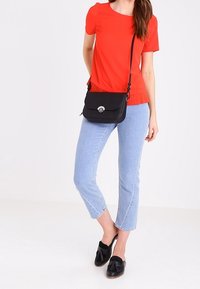 Černá crossbody taška s hladkým povrchem a zakřiveným klopou. Ozdobena stříbrným uzávěrem. Nošena s červeným topem a světle modrými džíny.
