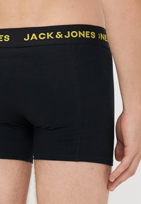 Mann trägt schwarze Boxershorts mit gelbem "JACK & JONES"-Logo am Bund, zeigt unteren Oberkörper und Oberschenkel.