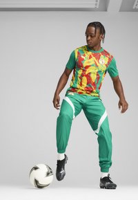 Ropa deportiva verde con un vibrante patrón floral en rojo, amarillo y verde, combinada con botas negras. Un balón de fútbol está situado cerca.