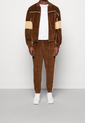 Muž v hnědém velurovém teplákovém obleku s béžovými pruhy na rukávech a kalhotách, v bílém tričku a bílých teniskách, stojící před jednobarevným pozadím.