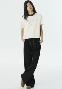 Crème crop top t-shirt met de tekst "UNIVERSE" en een bruine kraag, gecombineerd met losse zwarte broek en beige instap schoenen.