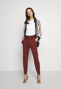 Cardigã com padrão floral e bordas azul-marinho, t-shirt branca e calças burgundy de cintura alta com cinto atado, complementadas por sandálias douradas de tiras.