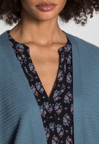 Světlomodrý pletený svetr přes tmavý vršek s květinovým vzorem a výstřihem do V. Obsahuje složité květinové detaily v červené a modré barvě na černém pozadí.