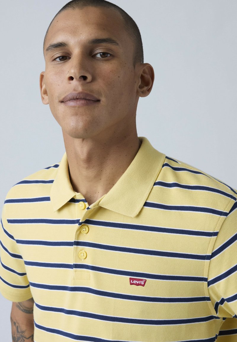 Homme aux cheveux courts portant un polo Levi's rayé jaune et bleu marine, debout devant un fond clair uni.