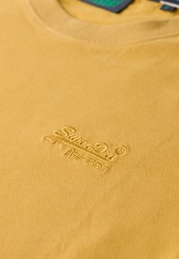 Camiseta de algodón amarilla con cuello redondo y un logotipo de la marca bordado en un amarillo más oscuro, ubicado en el centro delantero.