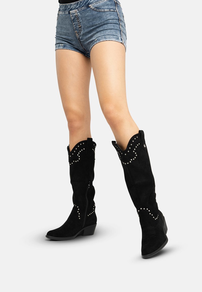 Schwarze kniehohe Stiefel aus Wildleder mit wellenförmigem oberen Rand, dekorativen silbernen Nieten und niedrigem Absatz; mit seitlichem Reißverschluss für einfaches An- und Ausziehen gestaltet.