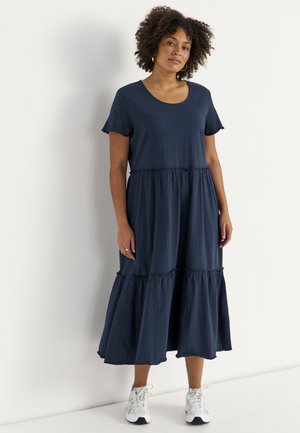 Vrouw met krullend haar, die een marineblauwe gelaagde midi-jurk en witte sneakers draagt, staand tegen een effen witte muur.