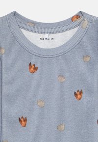Blau gerippter Stoff mit verstreutem braunem und beigem Blattmuster, das den runden Ausschnitt eines langen Kindershirts zeigt.