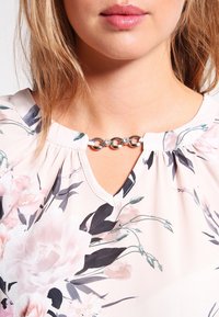 Blusa floreale con un tessuto morbido nelle tonalità di rosa e crema, dotata di un collo con apertura a chiave e dettagli in catena metallica al colletto.