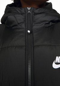 Chaleco acolchado negro con cuello alto, cremallera en la parte delantera y detalles cosidos. Presenta un logotipo de Nike en blanco en el pecho. Textura suave.