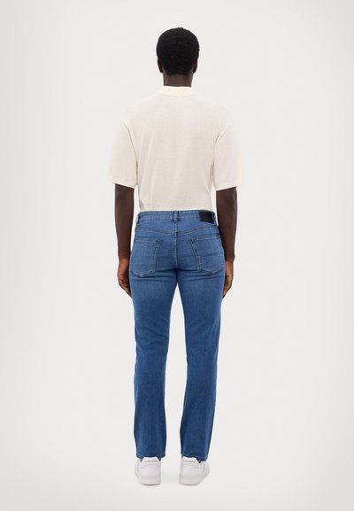 Meeste sinised denim püksid, millel on kitsas lõige ja sirged jalad, koos heleda värvi kootud polo särgiga. Omab kahte tagataskut ja denimitekstuuri.