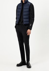 Mężczyzna ubrany w granatową pikowaną kamizelkę na czarną bluzę z długim rękawem, czarne dopasowane spodnie i czarne skórzane botki, stojący z rękami w kieszeniach.