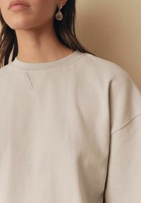 Jasno beżowa bluza z okrągłym dekoltem, opadającymi ramionami i subtelnym detalem w kształcie litery V. Połączona ze złotymi kolczykami w kształcie muszli.