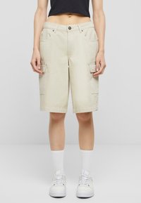 Beige cargo shorts med sidelommer, løs passform, liv med knapp og sydd kant. Brukt med hvite joggesko og sokker.