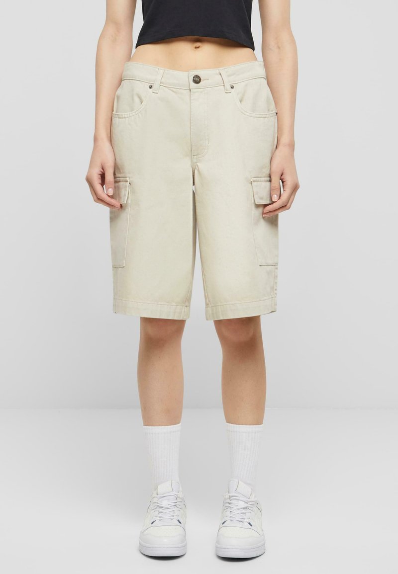 Beige cargo shorts med sidelommer, løs passform, liv med knapp og sydd kant. Brukt med hvite joggesko og sokker.