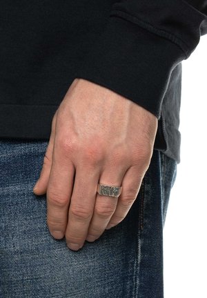 Silberner, gravierter Ring an einer Hand, getragen mit einem schwarzen Langarmshirt. Die Hand ruht auf blauen Jeans, die eine glatte, polierte Textur zeigen.