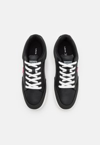 Baskets synthétiques noires avec des lacets blancs, des bouts arrondis, et une semelle blanche contrastante. Elles comportent une petite étiquette de marque rouge sur le côté.