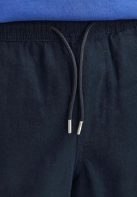 Gros plan sur un pantalon à taille élastique bleu marine avec des cordons noirs et des embouts argentés, porté avec une chemise bleue.