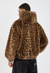 Bărbat îmbrăcat cu o jachetă cu glugă din blană artificială cu imprimeu de leopard maro și negru, cu tiv elastic, și pantaloni negri, arătat din spate pe un fundal alb.