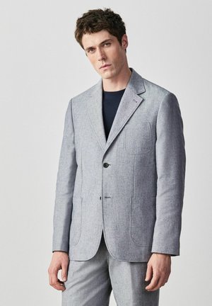 Junger Mann, der einen hellgrauen Blazer und passende Hose über einem schwarzen Hemd trägt, vor einem schlichten hellen Hintergrund stehend.