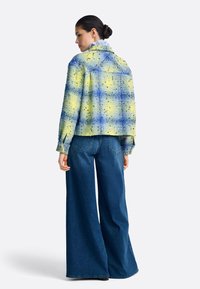 Rjavo modra karirasta jakna z teksturirano tkanino, nošena preko širokih kavbojk z razširjenimi hlačnicami. Ima gumbe na manšetah in krojeni ovratnik.