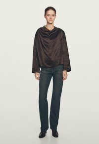 Donna che indossa un top lungo marrone scuro e morbido, jeans dritti blu scuro e stivaletti neri con tacco, in piedi contro uno sfondo neutro.