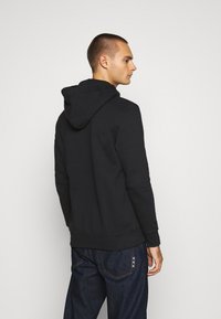 Felpa nera con cappuccio regolabile, polsini a costine e tessuto texturizzato. Indossata con jeans blu scuro caratterizzati da cuciture decorative.