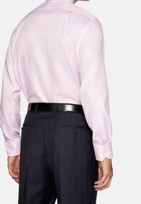 Man med en ljusrosa skjorta instoppad i mörka ränderade kostymbyxor och ett svart bälte, sett bakifrån.