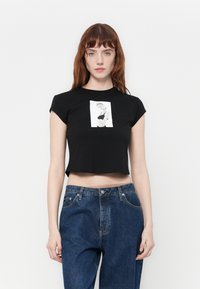 Zwarte cropped t-shirt met korte mouwen, voorzien van een centraal zwart-wit grafisch ontwerp. Gecombineerd met hoge blauwe denim jeans.