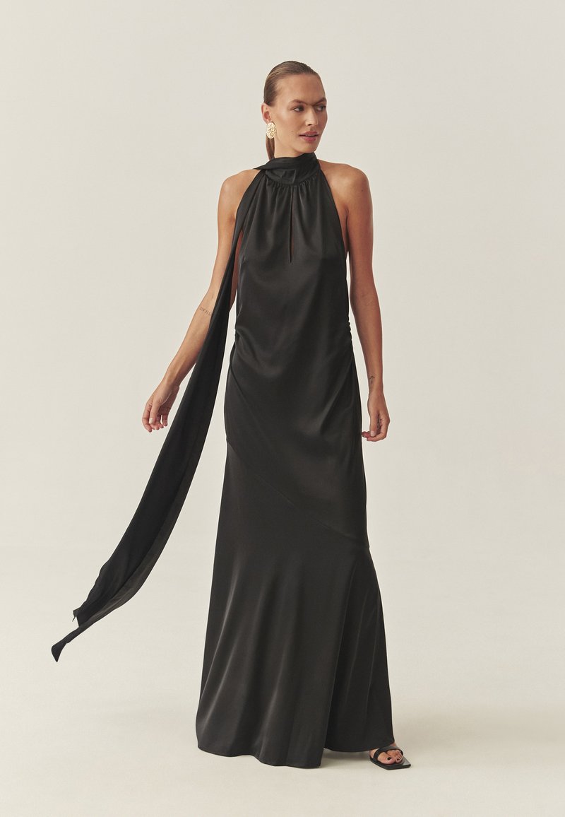 Vestido de raso negro con cuello halter y un detalle de capa drapeada, que presenta un cuello alto y una silueta fluida que se estrecha en el dobladillo.