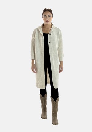 Mujer con una larga rebeca cremosa con botones, top y pantalones negros, y botas marrones con estampado hasta la mitad de la pantorrilla, de pie frente a un fondo liso.