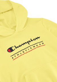 Felpa gialla realizzata in tessuto morbido, con un grande tascone frontale e un logo che recita "Champion ATHLETICWEAR" in testo nero e rosso.