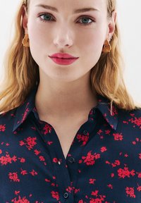 Kobieta z falującymi blond włosami, nosząca złote kwiatowe kolczyki i granatową koszulę w czerwoną kwiatową grafikę, patrząca prosto przed siebie.