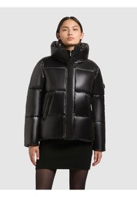 Melna pūšļa jaka ar augstu apkakli, spīdīgu apdari, kvadrātveida formu un pildītām daļām. Iekļauj rāvējslēdzēja aizdari un sānu kabatas.