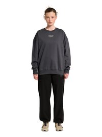 Kobieta stojąca, ubrana w ciemnoszarą oversize'ową bluzę z napisem "HALO", czarne luźne spodnie i beżowe trampki na białym tle.