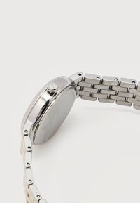 Ασημένιο μεταλλικό ρολόι χειρός με συνδεδεμένο λουράκι, η πλευρική κορώνα φέρει χαραγμένο το «ck», εμφανίζεται από την πλευρά και την πίσω όψη του ρολογιού πάνω σε λευκό φόντο.