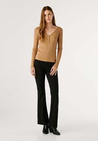 Lichtbruine ribbeltop met lange mouwen en een V-hals, voorzien van drie knopen, gecombineerd met zwarte flared broek en zwarte enkellaarsjes met hak.