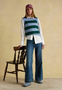 Megztinis be rankovių su mėlynomis ir žaliomis juostelėmis, baltas marškinis, mėlyni platėjančios džinsai ir pilki klumpes. Purpurinė kepurė ir medinė kėdė fone.