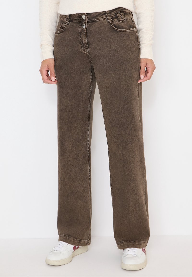 Jean marron à jambes larges avec taille à double bouton, porté avec des baskets blanches et un haut à manches longues clair.