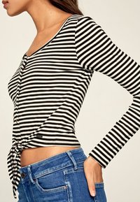 Svart och vit randig långärmad topp med scooped halsringning och knapppdetalj, knuten i midjan, tillsammans med blå jeans.