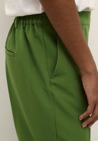Pantaloni leggeri verdi con vita elasticizzata, tasca laterale e texture liscia, mostrati di lato su uno sfondo neutro.