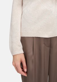 Jasnobeżowy sweter z teksturowanym wzorem i prążkowanymi mankietami, w połączeniu z brązowymi spodniami w kant. Skoncentrowano się na miękkim materiale i kontraście kolorystycznym.