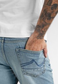 Jeans in denim azzurro chiaro con una tasca posteriore che mostra un design a punto ondulato di contrasto. Mano appoggiata nella tasca, con un tatuaggio visibile.