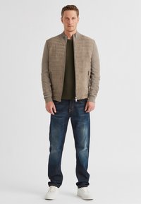 Beige gesteppte Bomberjacke mit einem gerippten Kragen, kombiniert mit einem dunkelgrünen strukturierten Hemd und blauen Jeans. Weiße Sneaker.