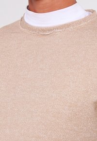 Beige svetr s texturovaným povrchem a kulatým výstřihem, vrstvený přes bílý rolák. Viditelné stehy zdobí límec.